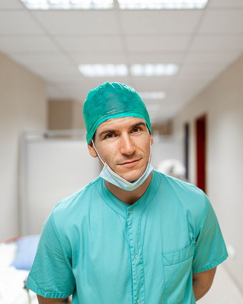 A Nurse Practitioner smiling wearing teal scrubs, cap and a mask at his chin, while standing in a hospital hallway.