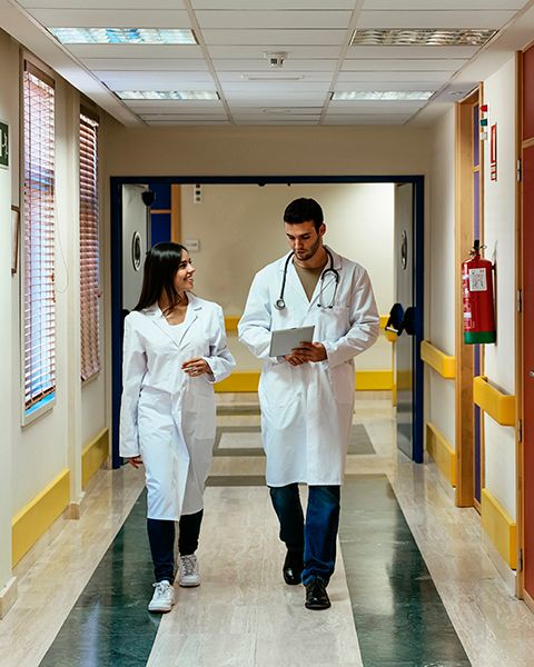 Two physicians walking down a hospital hallway, both in white coats, one a Hispanic woman, the other a Persian man.
