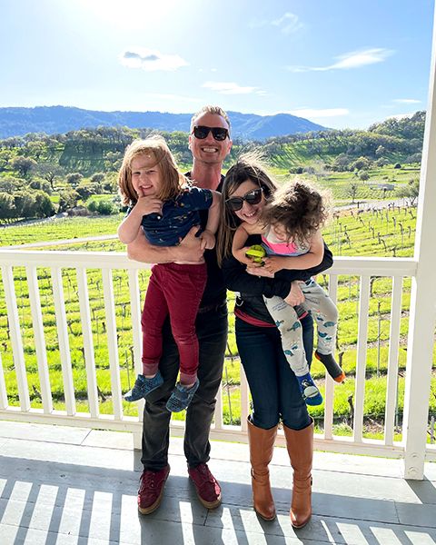 Dr. Dina Vaynberg, a white woman, standing next to her husband, a white man, smiling and hugging both of their toddler sons.