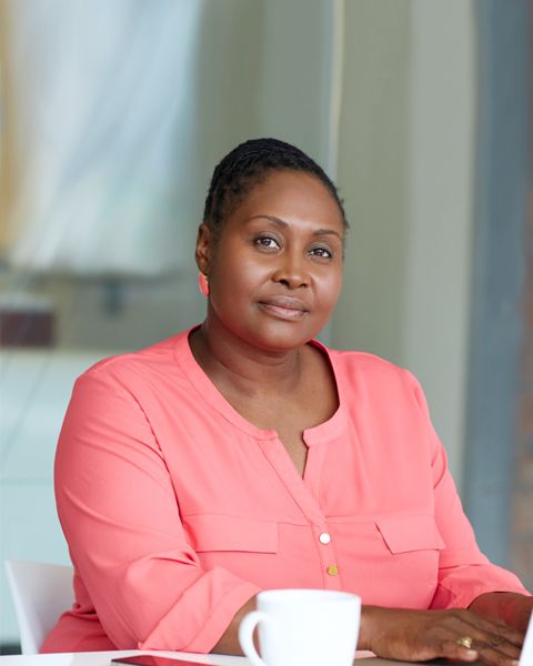 A hospital Executive, a Black woman, in a coral top and jewelry, smiling with a coffee cup next to her.