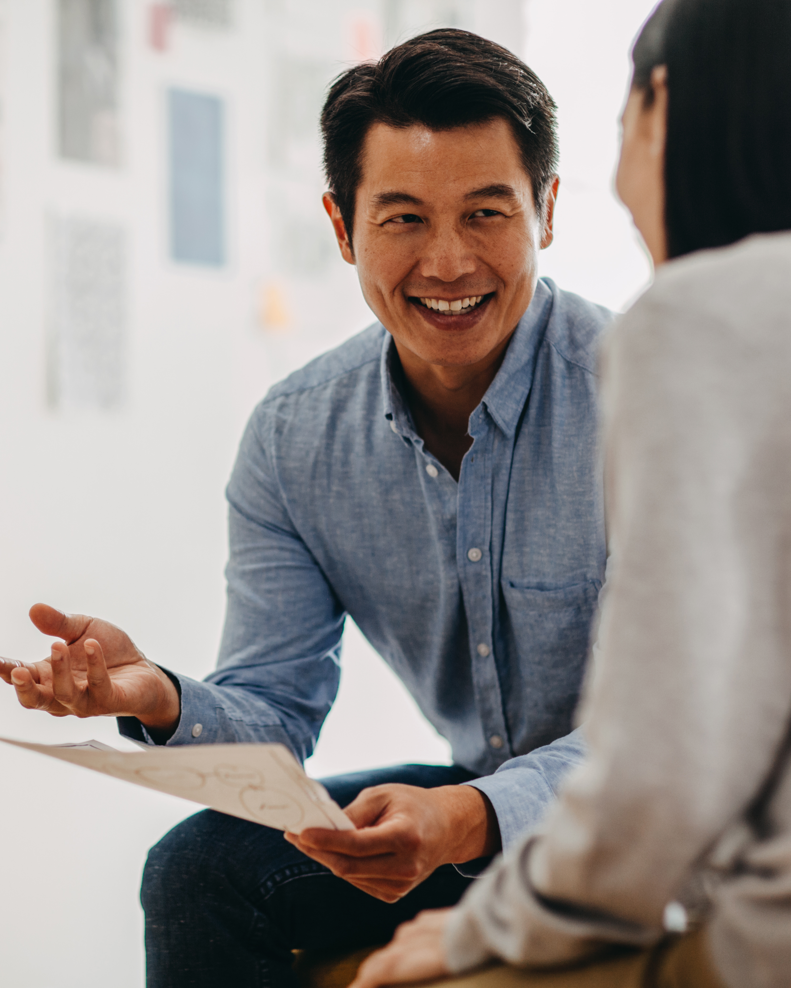 A physician, an Asian man, in a blue oxford shirt discussing a career change in medicine with a recruiter, an Asian woman.