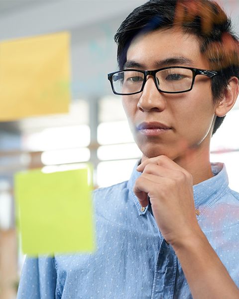 A physician, an Asian man, in blue oxford shirt reviewing two sticky notes. One has clinic written the other hospital.