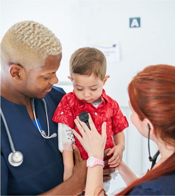 A physician, a Black woman, holding a toddler while another physician, a white woman, uses a stethoscope. 