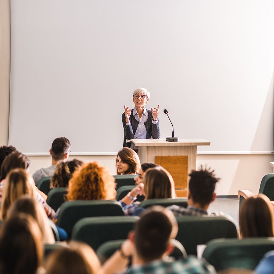 A Doctoral Faculty Member, a white woman with short white hair, teaching in front a class of students. 