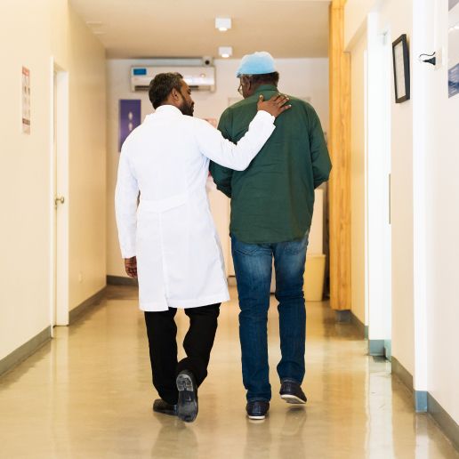 A physician with his arm over his patient's shoulder as they both walk down a hallway together, both are Black.