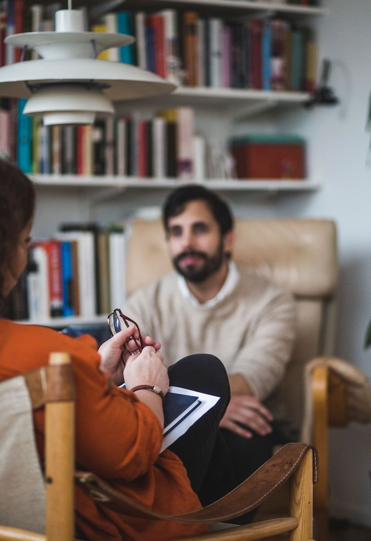 A Psychiatrist, a Hispanic woman, holding her glasses in hand, in session with her patient, a white man, in a beige sweater. 