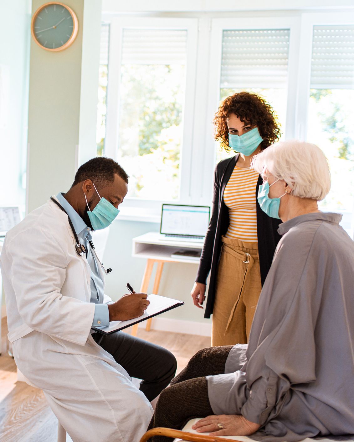 Physician, a Black man, in a white coat, with his patient, a senior white woman whose adult daughter stands nearby.